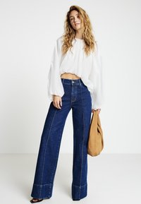 Jeans in denim scuro a gamba larga con cuciture a contrasto, abbinati a una blusa bianca corta. Una borsa color cuoio è tenuta nella mano destra.