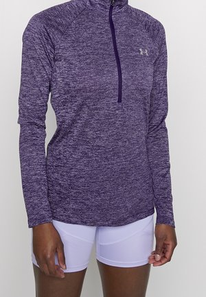 Lila melierter langärmeliger sportlicher Pullover mit Viertel-Reißverschluss, kombiniert mit weißen eng anliegenden Fahrradhosen an einer Person.