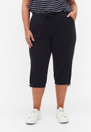 Pantaloni neri a vita alta con cordoncino, tasche laterali e polsini arricciati. Realizzati in un tessuto morbido con una texture liscia. Indossati con scarpe da ginnastica bianche.