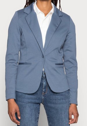 Kvinde iført en tætsiddende blå blazer over en hvid skjorte med krave og blå jeans, står med armene afslappet ned langs siderne.