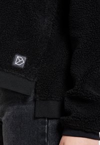 Giacca in pile nero con texture, dotata di polsini e orlo a costine, indossata sopra jeans scuri, con un piccolo ricamo sulla parte anteriore inferiore.