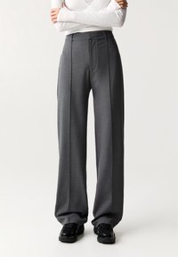 Pantalons larges gris en tissu texturé, avec des plis avant, une taille haute et des poches latérales, associés à des baskets noires.