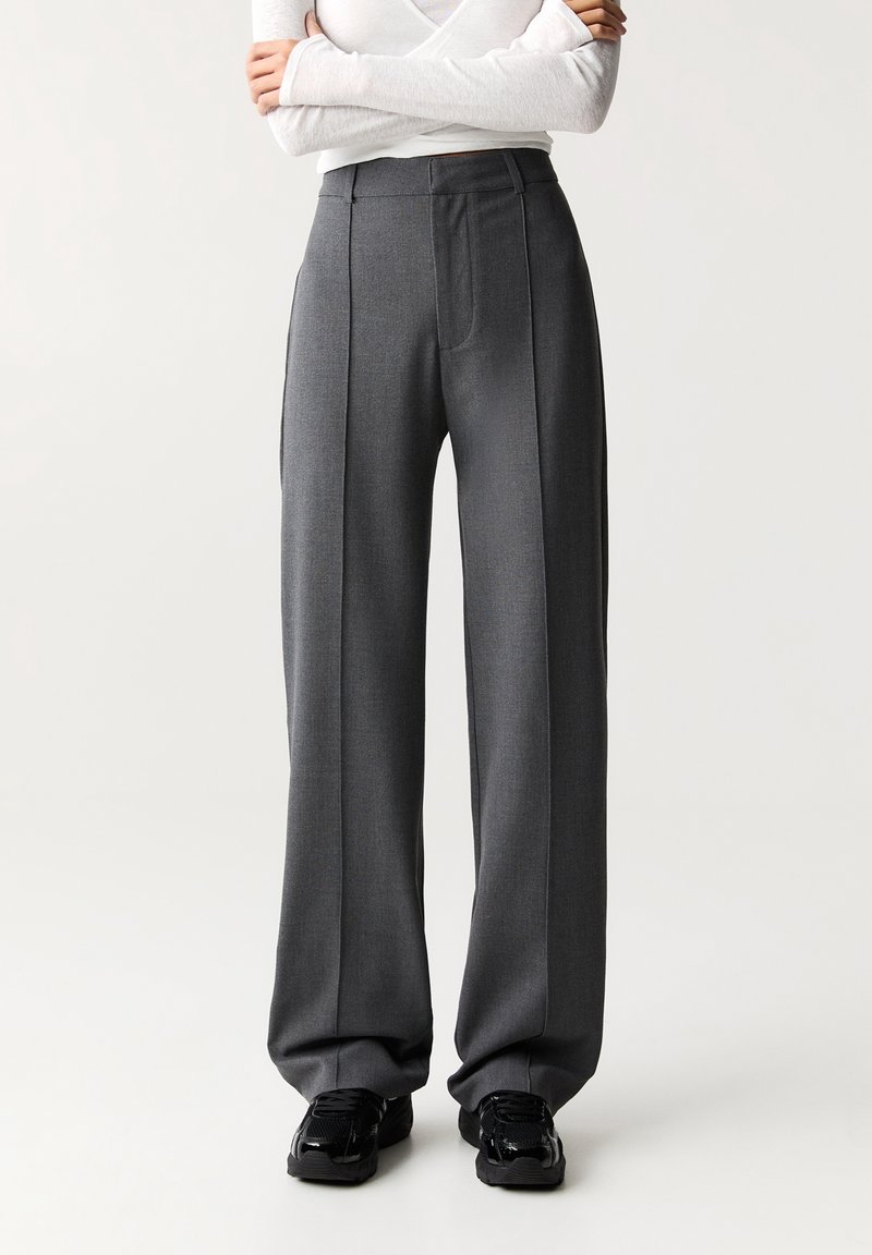 Pantalons larges gris en tissu texturé, avec des plis avant, une taille haute et des poches latérales, associés à des baskets noires.