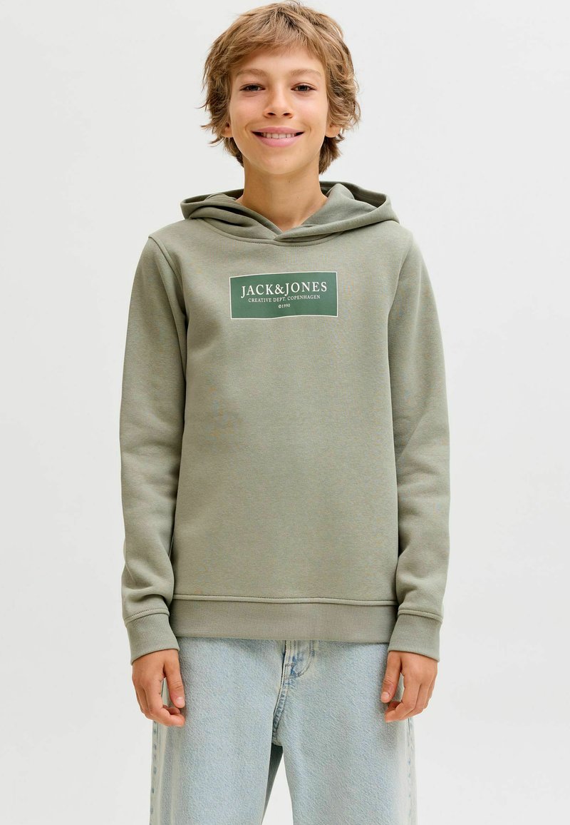 Žalias džemperis su kapišonu, su baltu logotipo pleistru ant krūtinės. Pagamintas iš minkštos medžiagos, su raštuotais rankogaliais ir apatine dalimi. Dėvimas su šviesiai mėlynomis džinsomis.