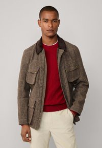 Bruine geruite jas met een donkere kraag, voorzien van twee voorzakken; gedragen over een rode kabeltrui en crèmekleurige broek. Verfijnde textuur.