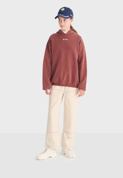 Jeune personne portant un sweat à capuche couleur rouille, un pantalon large crème avec des coutures rouges, des chaussures blanches épaisses et une casquette bleu marine avec un emblème.