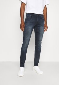 Jeans azul oscuro de corte ajustado con textura deslavada, diseño de cinco bolsillos y costuras en contraste, combinados con zapatillas blancas y una camiseta blanca holgada.