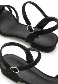 Schwarze Sandale mit flacher Sohle, geflochtenen Riemen und einer silbernen Schnalle zur Anpassung. Hergestellt aus Wildleder mit einer glatten Innenseite.