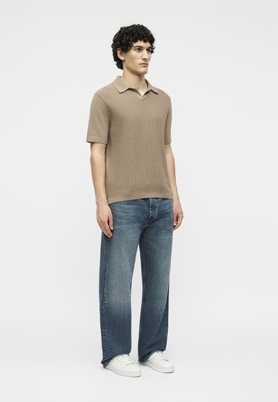 Άνδρας που στέκεται φορώντας μπεζ κοντομάνικο ριπ κοντομάνικο πόλο, μπλε τζιν με φαρδιά πόδια και λευκά αθλητικά σε απλό ανοιχτό φόντο.