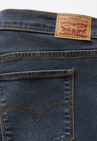 Tamsiai mėlyni džinsai iš džinsinio audinio su odine nugara lopu, kuriame raudonos spalvos tekstas ir dizainas. Išraiškingos auksinės siūlės ir išlenktų nugaros kišenių siūlės.