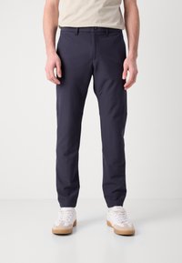 Marinblå byxor med smal passform, framfickor och en slät textur, tillsammans med vita sneakers och en beige skjorta.