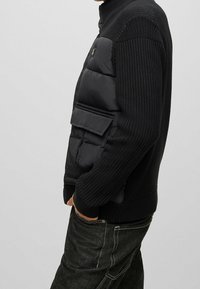 Maglione nero lavorato a maglia con una parte anteriore imbottita, caratterizzato da un colletto a zip, una grande tasca frontale e maniche testurizzate. Abbinato a jeans scuri.