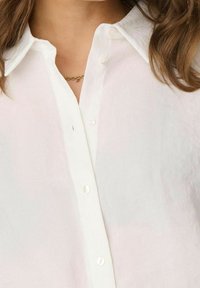 Chemise blanche boutonnée avec un col classique, un tissu léger et des boutons visibles. La texture semble lisse et légèrement transparente.