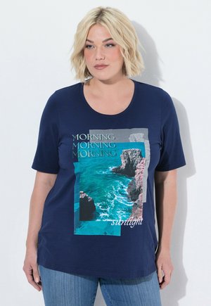 Lyshåret kvinde iført navyblå t-shirt med grafik af havklipper og bølger, tekst "MORNING" gentaget og "sunlight", blå jeans, stående mod en hvid væg.