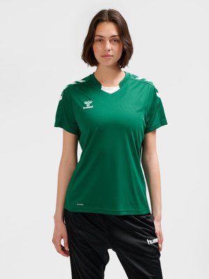 Vrouw die een groen sportshirt met wit logo en zwarte sportbroek draagt, staand tegen een effen lichte achtergrond.