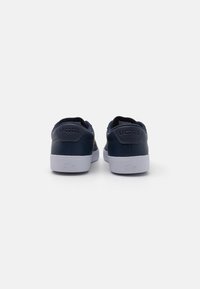 Navy leather sneakers with white rubber soles, featuring a textured finish and embossed "LACOSTE" logo on the back. Round toe design.