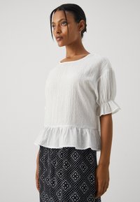 Bluză albă texturată cu un tiv peplum, guler rotund și mâneci bufante. Asortată cu o fustă neagră cu un model în forme de diamant.