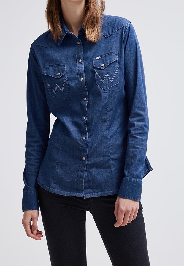 Femme portant une chemise en denim bleu foncé ajustée à manches longues avec des poches à rabat boutonnées et un pantalon noir, debout devant un fond uni.