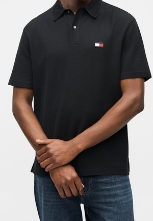 Mann trägt ein schwarzes kurzärmeliges Poloshirt mit einem kleinen roten, weißen und blauen rechteckigen Logo auf der Brust und blaue Jeans, Hände verschränkt.
