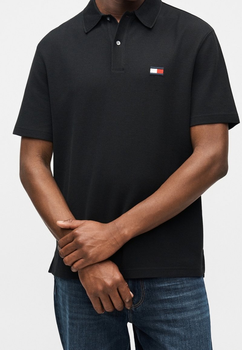 Homme portant un polo noir à manches courtes avec un petit logo rectangulaire rouge, blanc et bleu sur la poitrine et un jean bleu, les bras croisés.