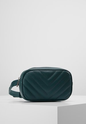 Sac banane en cuir texturé vert foncé avec coutures chevrons, une fermeture éclair argentée et une sangle réglable sur une surface blanche.