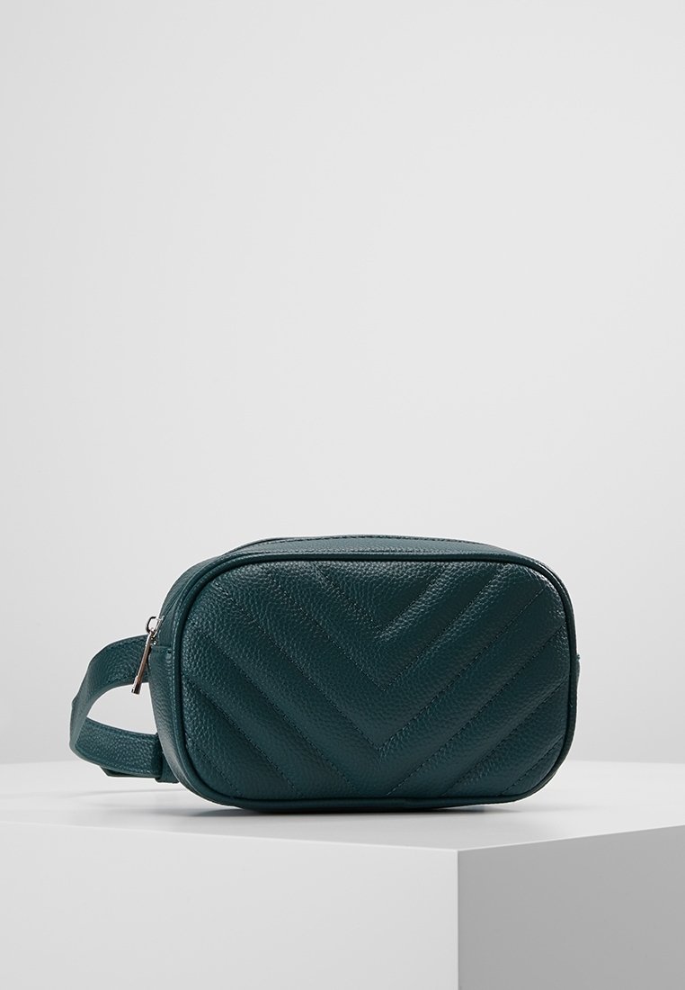 Sac banane en cuir texturé vert foncé avec coutures chevrons, une fermeture éclair argentée et une sangle réglable sur une surface blanche.