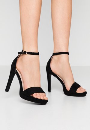 Frau trägt schwarze Sandalen mit hohen Absätzen, Knöchelriemen und offenen Zehen, steht auf einem hellgrauen Boden vor einem schlichten Hintergrund.