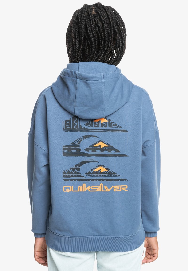 Personne avec des cheveux tressés portant un sweat à capuche bleu avec trois motifs de vagues stylisées et un logo Quiksilver orange dans le dos.