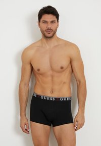 Black boxer briefs with a thick waistband featuring the "GUESS" logo in white and red, made from a smooth, stretchy material.
