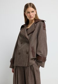 Giacca doppiopetto marrone con texture, dettagli in pelle sulle spalle e sui polsini, dotata di un ampio colletto e di una vestibilità rilassata.