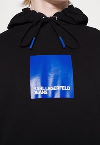 Sudadera negra con un logo azul cuadrado que lleva el texto "KARL LAGERFELD JEANS" en blanco. Cuerdas con puntas azules en la capucha.
