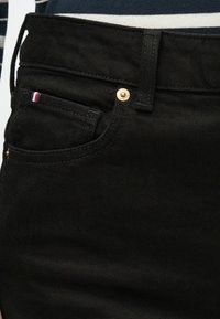 Jeans de ganga pretos com um bolso frontal que exibe uma faixa vermelha, branca e azul. Detalhe de botão dourado na cintura e tecido texturizado.