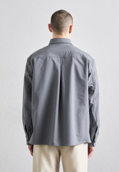 Gray long-sleeve shirt with a back pleat, straight cut, buttoned cuffs, and a collar. Made from a smooth fabric with minimal detailing.