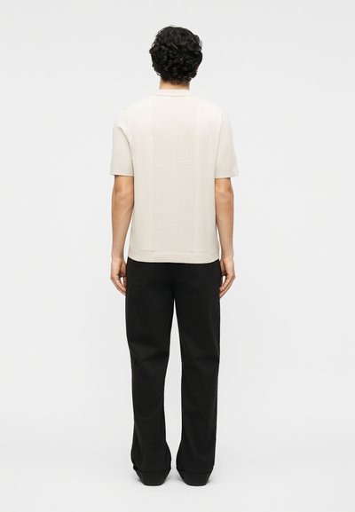 Άνδρας με σγουρά μαύρα μαλλιά, φοράει μπεζ πουκάμισο με κοντά μανίκια και υφασμένη πλάτη, καθώς και μαύρο παντελόνι με φαρδιές μπατζάκια, στέκεται με πλάτη προς τα εμπρός.