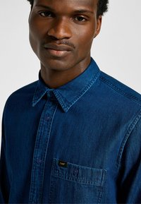 Granatowa koszula z denimu z kołnierzykiem zapinanym na guziki, kieszonką na piersi oraz metką marki. Gładka tekstura, dopasowany krój.