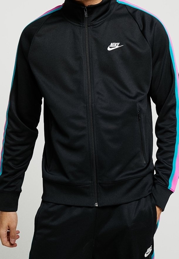 Man wearing black Nike tracksuit with pink and blue stripes on sleeves and pants, standing with one hand in pocket.