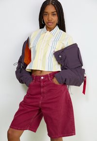 Camicia a maniche corte, con bottoni, a righe gialle e blu, abbinata a pantaloni corti bordeaux a vita alta. Una giacca viola scuro pende dalle spalle.
