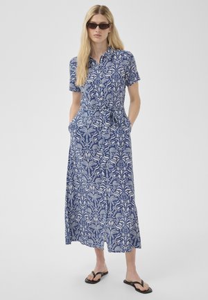 Femme portant une robe maxi à manches courtes boutonnée avec ceinture, à motif bleu et blanc, lunettes de soleil noires et tongs noires, mains dans les poches.