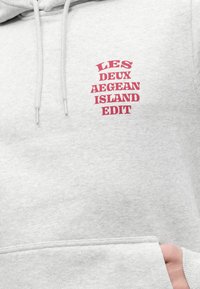 Φωτεινή γκρι μπλούζα με κόκκινο κείμενο "LES DEUX AEGEAN ISLAND EDIT" στο στήθος, φορεμένη με το χέρι εν μέρει στην μπροστινή τσέπη.