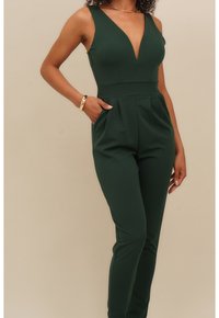 Kvinna iklädd en ärmlös mörkgrön jumpsuit med djup V-halsringning, guldarmband och halsband, stående med en hand i fickan.