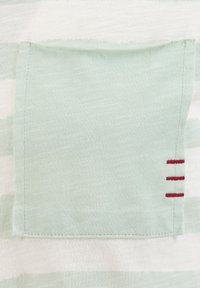 Tasca in tessuto verde chiaro cucita su un capo bianco a righe verde chiaro con tre brevi punti orizzontali rossi sul lato destro.