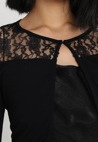 Černý kardigan s krajkovými detaily na ramenou, zapínáním na klíček a měkkou texturou, nošený přes tmavý top.