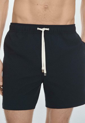 Muž nosící černé texturované plavky s bílým šňůrkováním, stojící před jednoduchým světlým pozadím.