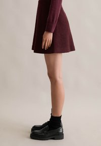 Bordowa, krótka sukienka typu skater z teksturowanej tkaniny z długimi rękawami. Połączona z czarnymi loafersami i skarpetkami do kostek. Widok z boku.