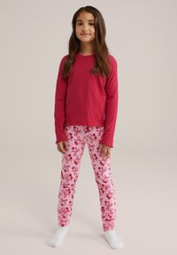 Piros, hosszú ujjú ing, fodros széllel, rózsaszín leggings-szel párosítva, amely sötétebb rózsaszín virágmintával díszített. Fehér zoknik.