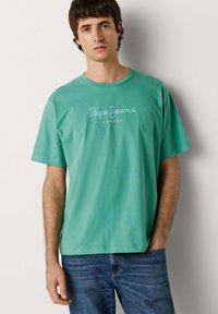 Ung mand iført en teal Pepe Jeans London t-shirt og blå jeans, stående mod en ensfarvet lys baggrund med den ene hånd i lommen.