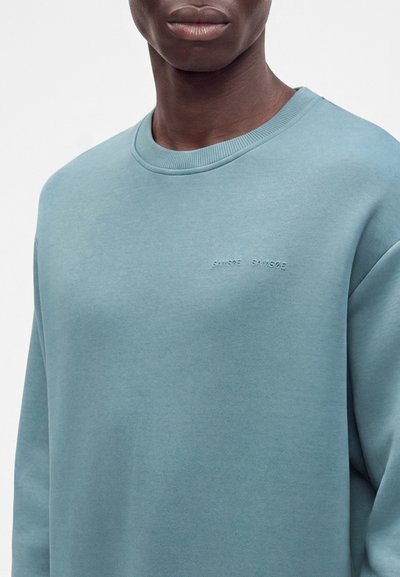 Gros plan d'une personne portant un sweat-shirt bleu clair avec un texte en relief subtil sur la poitrine indiquant "SAMSOE SAMSOE."