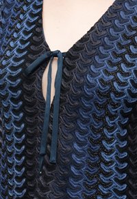 Κοντινό πλάνο μπλε και μαύρου πλεκτού ρούχου με κυματιστό σχέδιο δεμένο στον λαιμό, αφήνοντας να φανεί το δέρμα κάτω από το άνοιγμα.