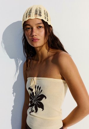 Young woman with long hair wears a knitted cream tube top with black floral design and a matching crochet beanie, standing against white wall.