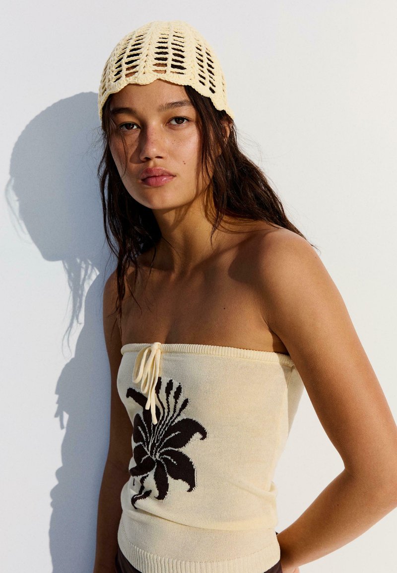 Jeune femme aux longs cheveux porte un haut tube en tricot crème avec un motif floral noir et un bonnet au crochet assorti, debout contre un mur blanc.